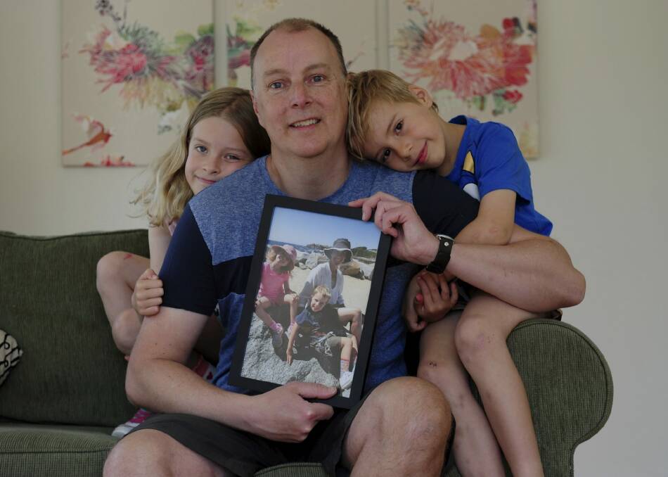 Trevor Hickman at his home in Curtin with his two children Audrey, 8, and Elijah, 6. He is holding a photograph of his wife, Amy, who died of breast cancer last year. Photo: Graham Tidy