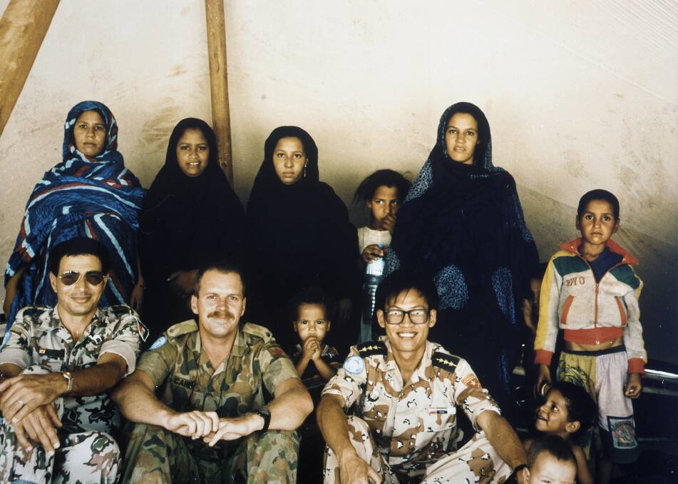 Group portrait of United Nations Military Observers (UNMOs) from MINURSO (United Nations Mission for the Referendum in Western Sahara) with Polisario wives and local children. Major Carey is seated second from left. Photo: Australian War Memorial